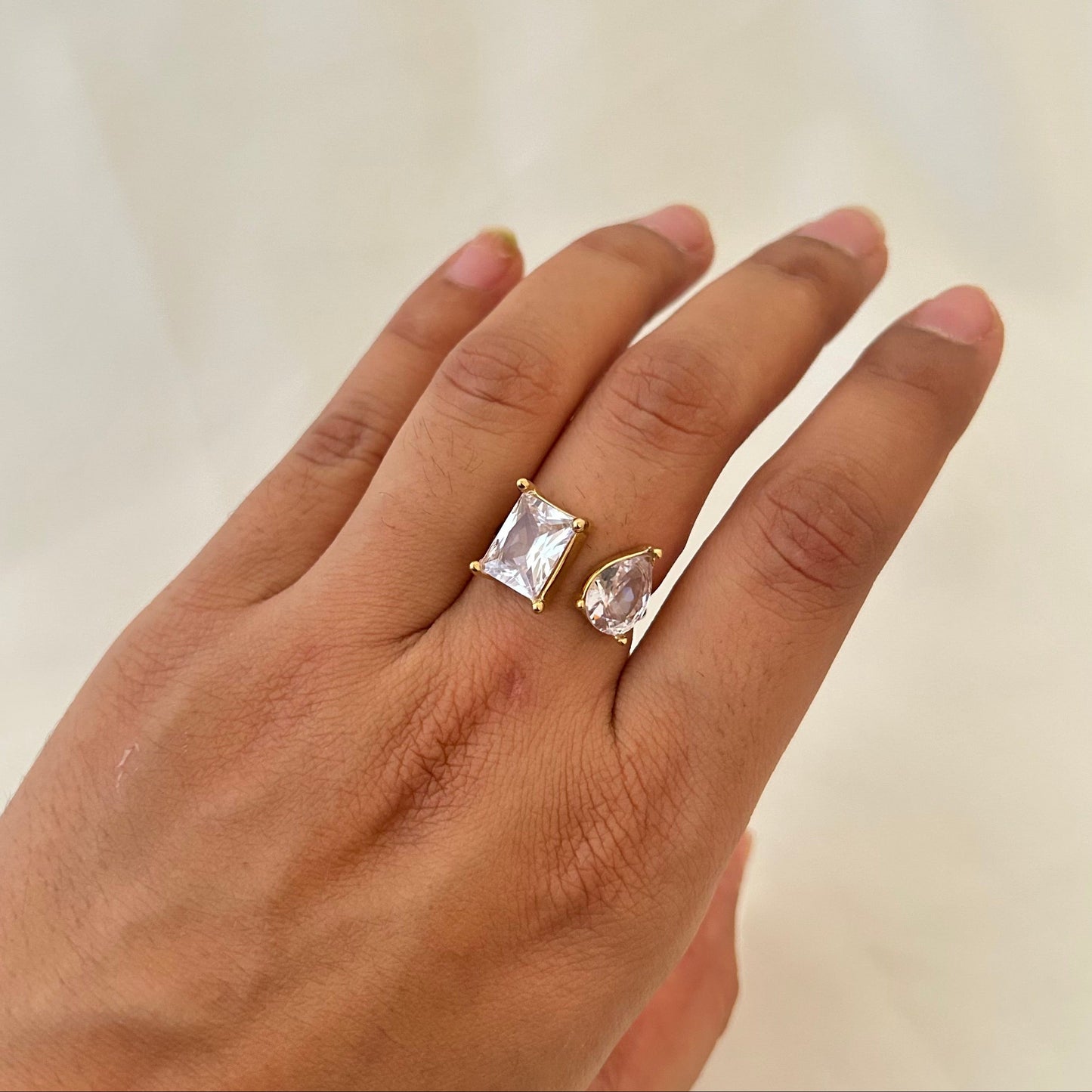 Hand wearing a gold open ring with two clear gemstones, one rectangular and one teardrop-shaped.