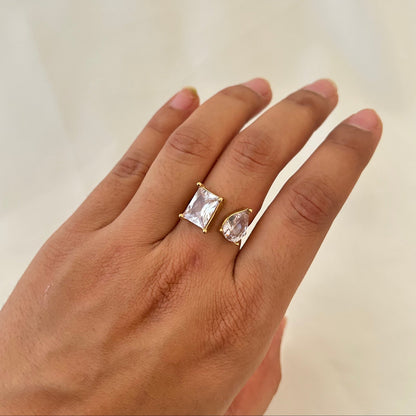 Hand wearing a gold open ring with two clear gemstones, one rectangular and one teardrop-shaped.