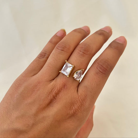 Hand wearing a gold open ring with two clear gemstones, one rectangular and one teardrop-shaped.